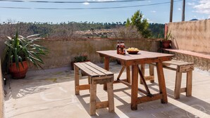 Et træbord udendørs med bænke, plantekrukker og flasker med øl på en tagterrasse med udsigt over landskabet.