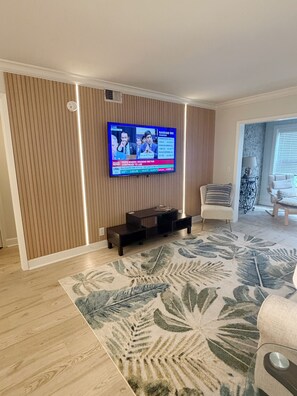 Un salon avec un téléviseur à écran plat fixé au mur, un meuble de rangement sombre en dessous, un tapis à motifs et un canapé de couleur claire.