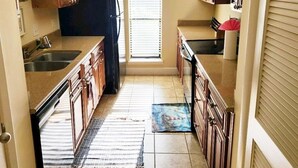 Una cocina con armarios de madera, un refrigerador negro, una ventana con persianas y piso de baldosas.