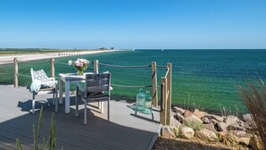 A wooden deck with a table and chairs, overlooking a beach and the ocean. - Luxus Beachvilla "the View Plus" (Ostseeresort Olpenitz)