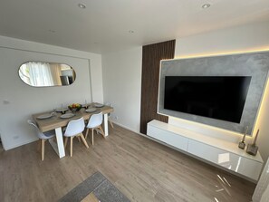 Un espace repas moderne avec un grand téléviseur, une table à manger en bois, des chaises blanches et un miroir.