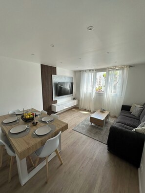A modern living room with a dining area, a flat-screen TV mounted on the wall, and a sofa with a view to the outdoors. - Spacious 3-bedroom near Paris - Private parking (Aubervilliers)