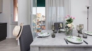 Ein Essbereich mit einem Tisch für zwei Personen, der mit Geschirr mit grünem Blattmuster, einer Blumenvase und Blick auf die Stadt durch ein Fenster ausgestattet ist.