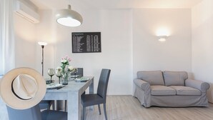 Ein modernes Wohnzimmer mit Essbereich, einem grauen Sofa, einem Esstisch mit Stühlen, einem Hut an der Wand und einer Abfahrtsanzeige.