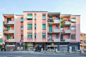 Un immeuble résidentiel à plusieurs étages, doté de balcons, avec une moto garée devant et des boutiques au rez-de-chaussée.