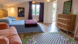 A bedroom with two beds, a large window with curtains, a door leading outside, a painting on the wall, and a wicker cabinet. - Rustic Barn Retreat in North Dorset (Gillingham)