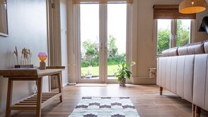 A spacious room with a wooden floor, a patterned rug, a sofa, a table with decorative items, and a view through a glass door to the outdoors. - Rustic Barn Retreat in North Dorset (Gillingham)