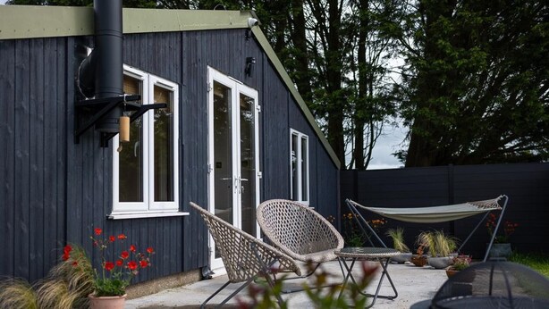 A house with a dark exterior, white-framed windows, and a door. There are two wicker chairs and a hammock outside. - Rustic Barn Retreat in North Dorset (Gillingham)