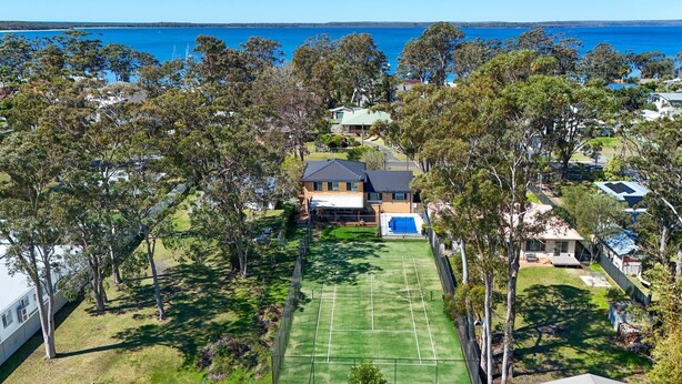 A residential area with a house featuring a tennis court, surrounded by trees and a view of the sea. - Anglesey - Pet Friendly with Pool - 4 Mins Walk to Beach (Callala Bay)