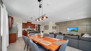 A modern living room with a wooden dining table, grey sofas, and a flat-screen TV mounted on the wall.