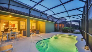Une piscine extérieure dotée d’une structure en verre, un espace repas avec mobilier d’extérieur, et une cuisine visible à travers le verre.