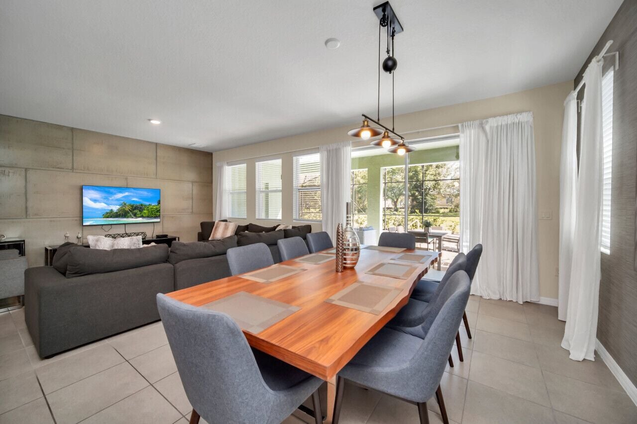 A modern living room with a large wooden dining table, grey sofas, and a TV mounted on the wall.