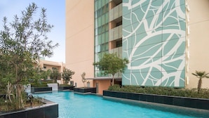 A swimming pool with a wooden deck, surrounded by greenery and modern buildings with a geometric facade. - Silkhaus AL Sana 2 - AL Raha (Abu Dhabi)