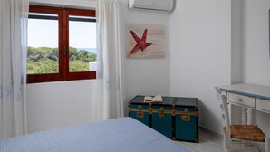 A bedroom with a bed, a window with curtains, a wall-mounted air conditioner, a wooden desk, a chair, and a blue trunk with a book. - Casa Dell Asfodelo by Wonderful Italy (Budoni)