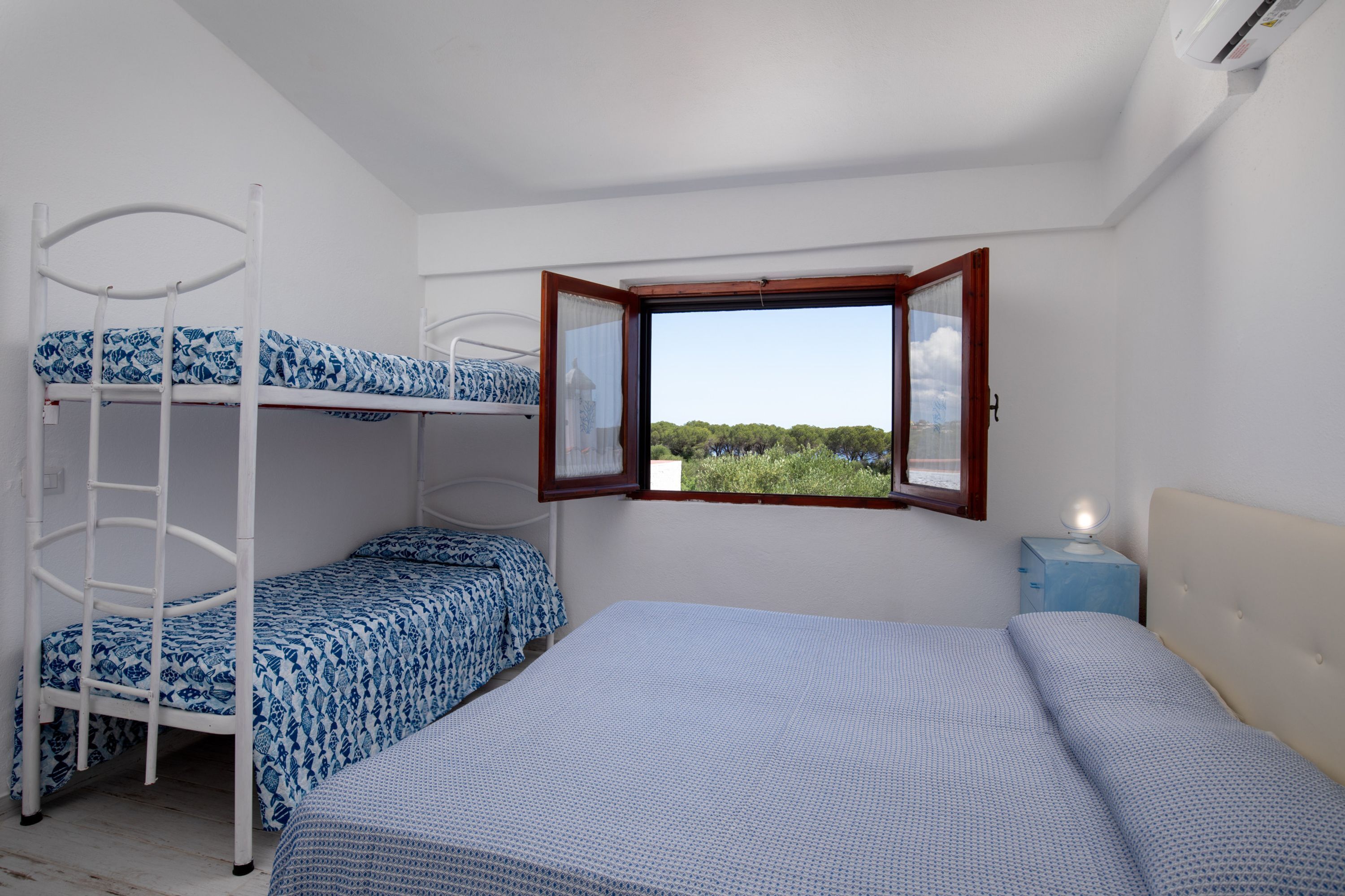 A room with bunk beds, a window with an open shutter, and a bedside table with a lamp.