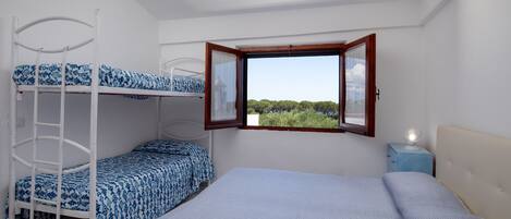 A room with bunk beds, a window with an open shutter, and a bedside table with a lamp.