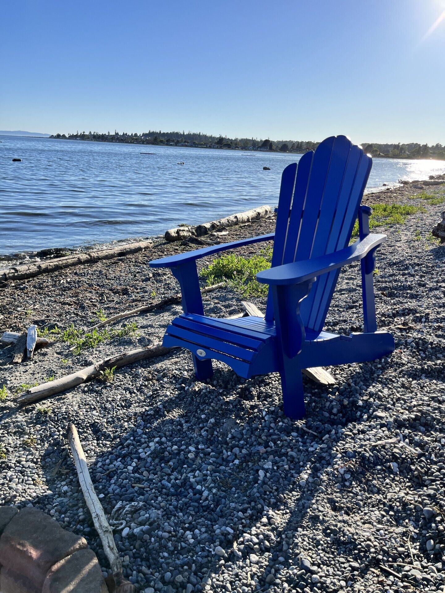 A blue Adirondack chair on a pebbled beach with a view of a calm lake and distant shoreline.