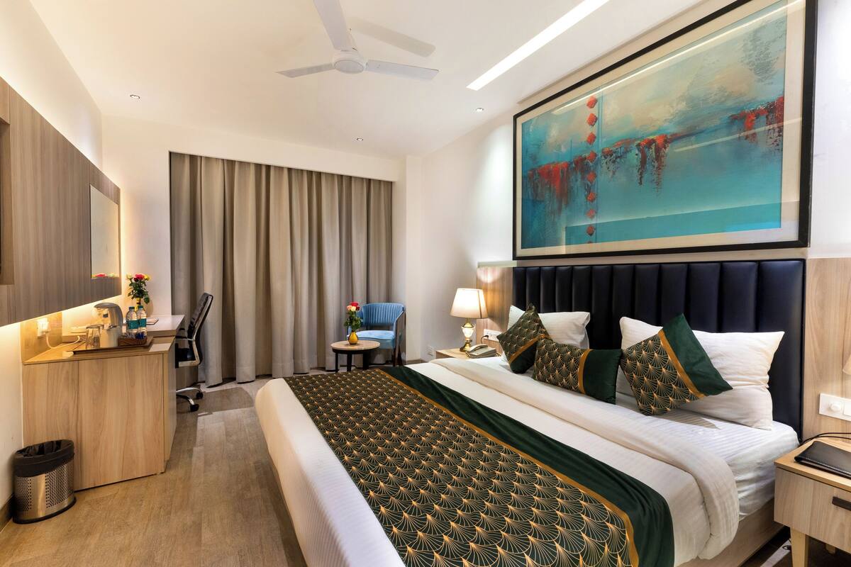A modern hotel room with a large bed, a desk, a chair, a ceiling fan, and a painting on the wall.