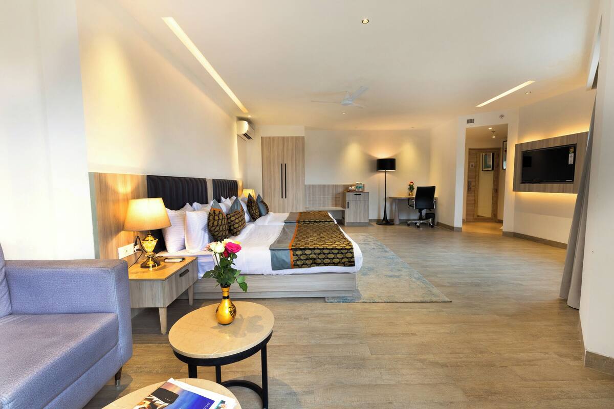 A modern hotel room with a sofa, a bed with a patterned bedspread, a small round table with a vase of flowers, a desk with a lamp, and a TV mounted on the wall.