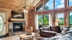 A cozy cabin interior with a stone fireplace, leather sofa, and large windows offering a view of the forest. - Cozy Private Cabin with Hot Tub & Mountain Views (Sevierville)