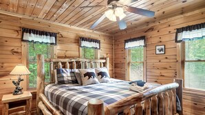 A log cabin bedroom with a wooden bed, a ceiling fan, and a window with a view of greenery. - Cozy Private Cabin with Hot Tub & Mountain Views (Sevierville)