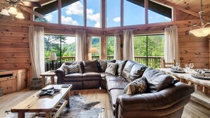 A spacious living room with a large L-shaped leather sofa, wooden coffee table, and a dining table set for four. Large windows provide a view of the outdoors. - Cozy Private Cabin with Hot Tub & Mountain Views (Sevierville)