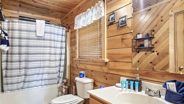 A wooden-paneled bathroom with a striped shower curtain, a toilet, a sink with toiletries, and a window with blinds. - Cozy Private Cabin with Hot Tub & Mountain Views (Sevierville)