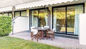 Eine Terrasse mit Tisch und Stühlen, einem Blumentopf und Blick durch eine Glastür in einen Raum mit Bett und Vorhängen.