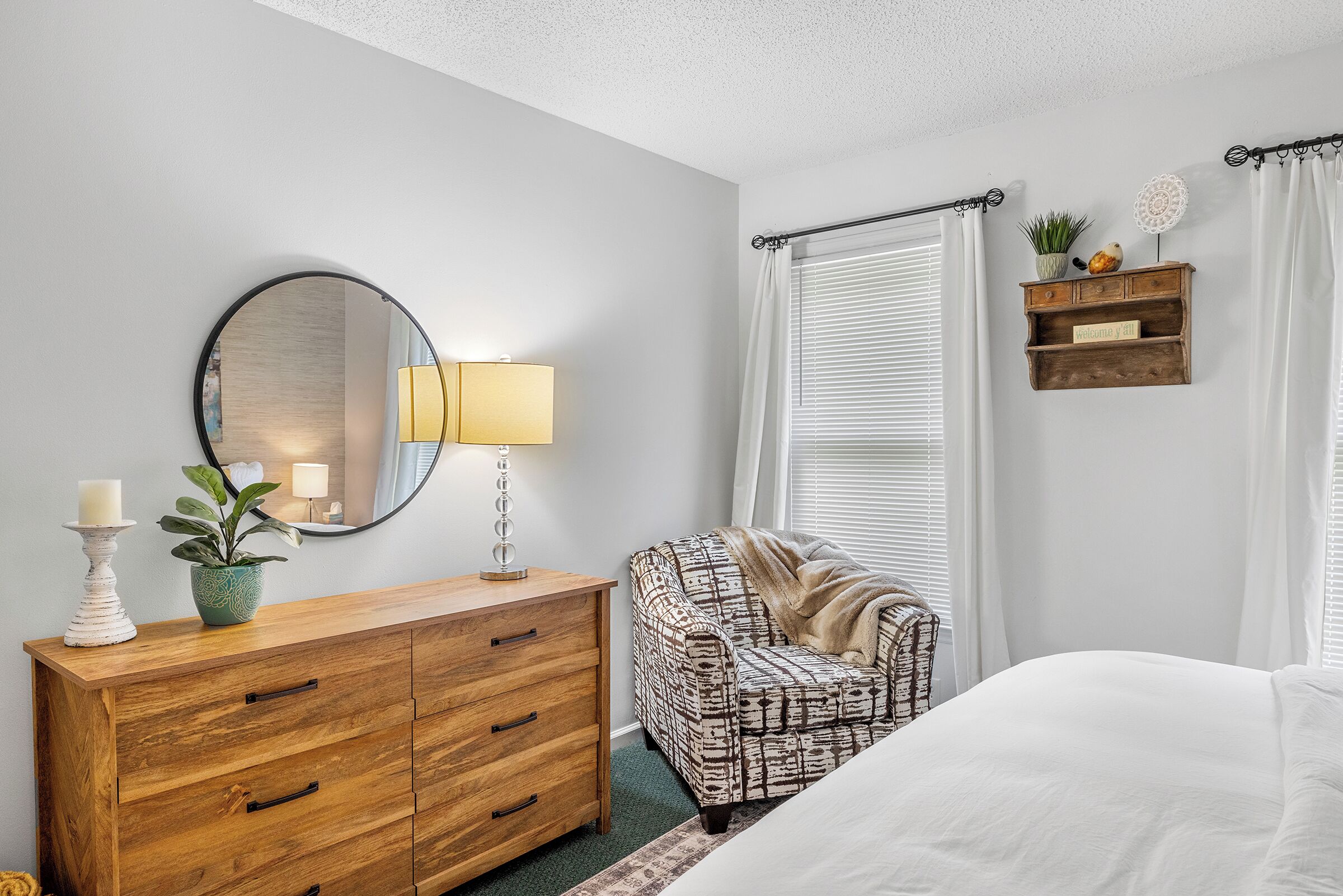 A bedroom with a wooden dresser, a patterned armchair, a bed, a mirror, a lamp, and a shelf with decorative items.