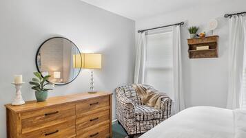 A bedroom with a wooden dresser, a patterned armchair, a bed, a mirror, a lamp, and a shelf with decorative items.