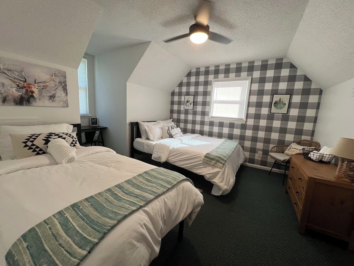 A bedroom with two beds, a ceiling fan, a window with blinds, and a checkered wall pattern.