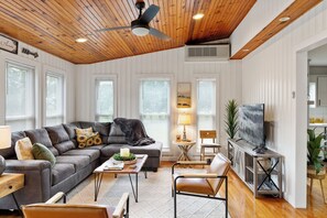 Un salon avec un plafond en bois, un canapé d’angle gris, une table basse, un meuble pour la télévision et un coin repas avec une chaise et une table.