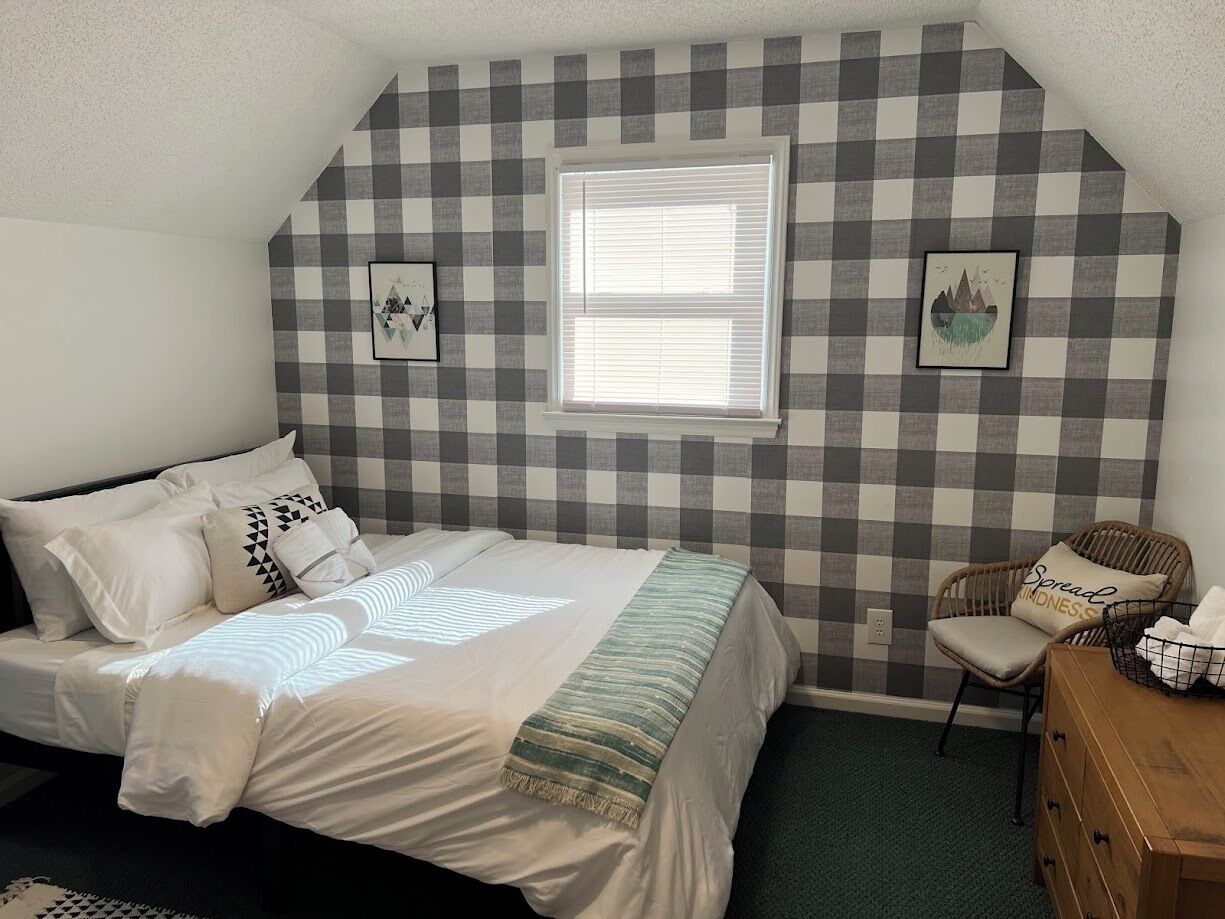 A bedroom with a checkered wall, a bed with white bedding, a window, a chair, and a wooden dresser.