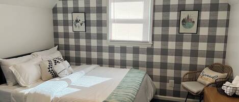 A bedroom with a checkered wall, a bed with white bedding, a window, a chair, and a wooden dresser.