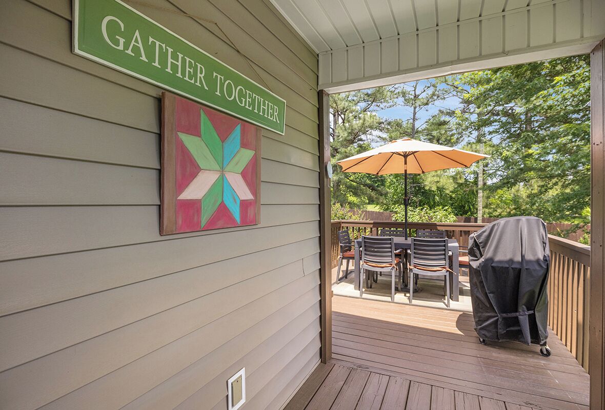 A covered outdoor area with a sign that reads "GATHER TOGETHER," a colorful geometric artwork, outdoor furniture, and a grill on a deck.