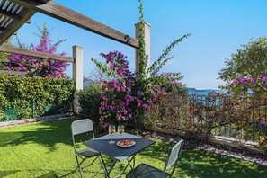Een terras met een tafel, stoelen, een pergola en bloeiende planten.