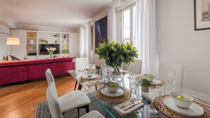 Uma sala de jantar moderna com uma mesa de vidro posta para dois, cercada por cadeiras brancas. Um sofá vermelho e um armário branco com objetos decorativos são visíveis ao fundo.