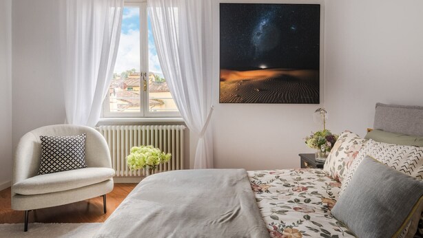 Quarto com cama, cadeira, pintura de céu noturno, janela com cortinas e radiador.