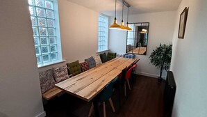 Un coin repas avec une table en bois, plusieurs chaises et un banc avec des coussins. De grandes fenêtres et un miroir ornent le mur.