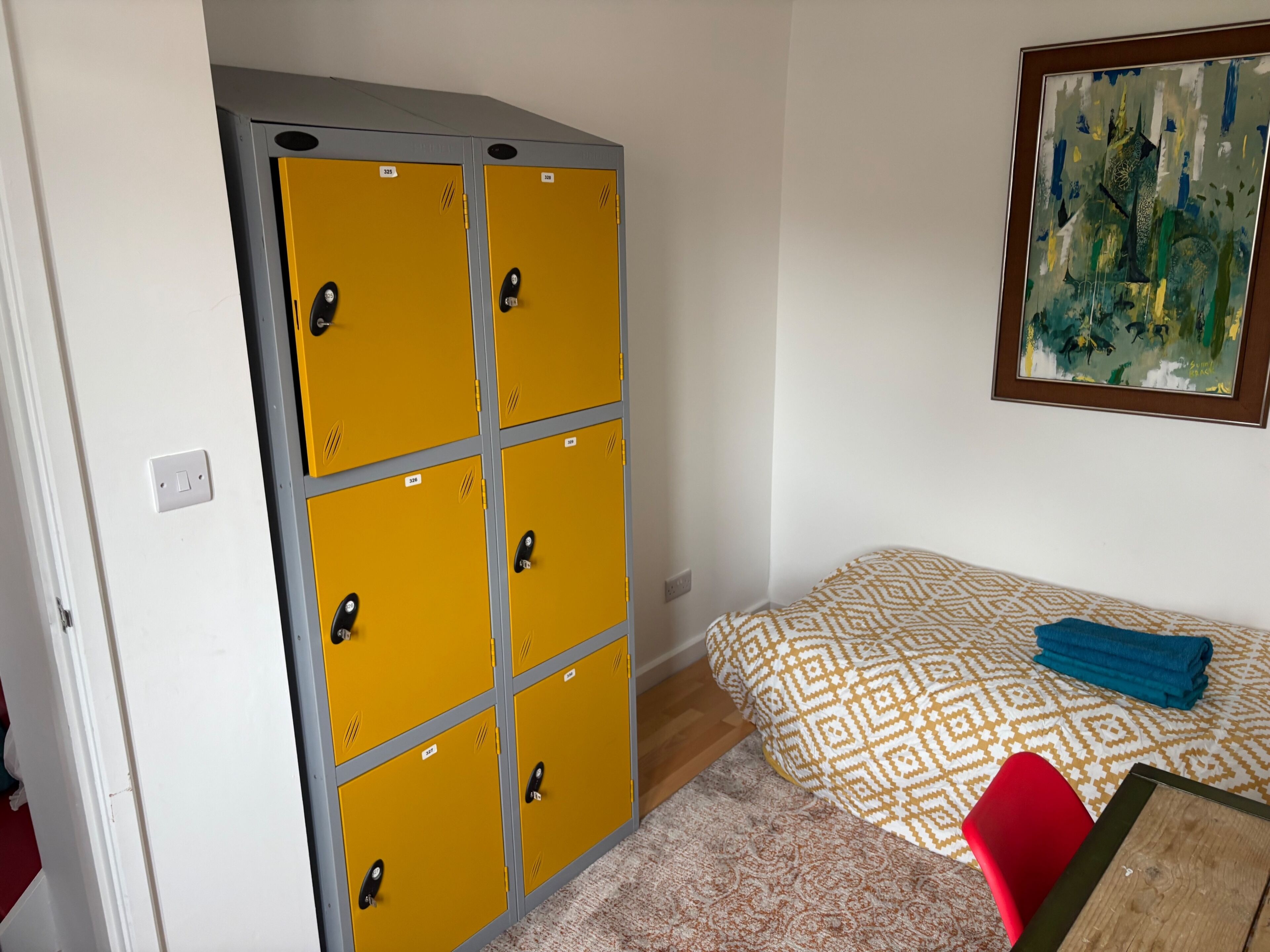 A room with a large yellow storage unit, a patterned bed, a blue towel, and a red chair.