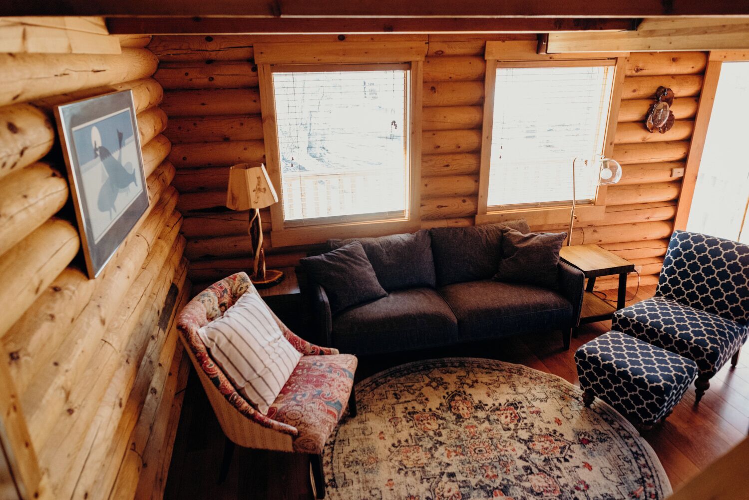 Acogedor interior de cabaña de madera con sofá, sillón, alfombra estampada y una pequeña mesa.