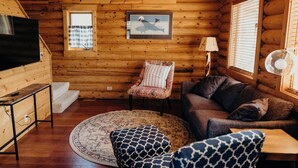 Ein gemütliches Blockhaus-Interieur mit Fernseher, einem gemusterten Sessel, einer Couch und einem runden Teppich.