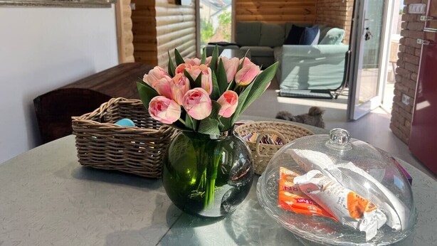 Een knus ingerichte kamer met houten details, een eettafel met een vaas roze bloemen en een vitrine met etenswaren.