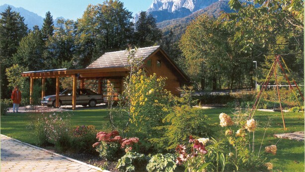 A wooden cabin with a car parked in front, surrounded by a garden with various plants and flowers, and a mountain range in the background. - Apartments Jakelj (Kranjska Gora)