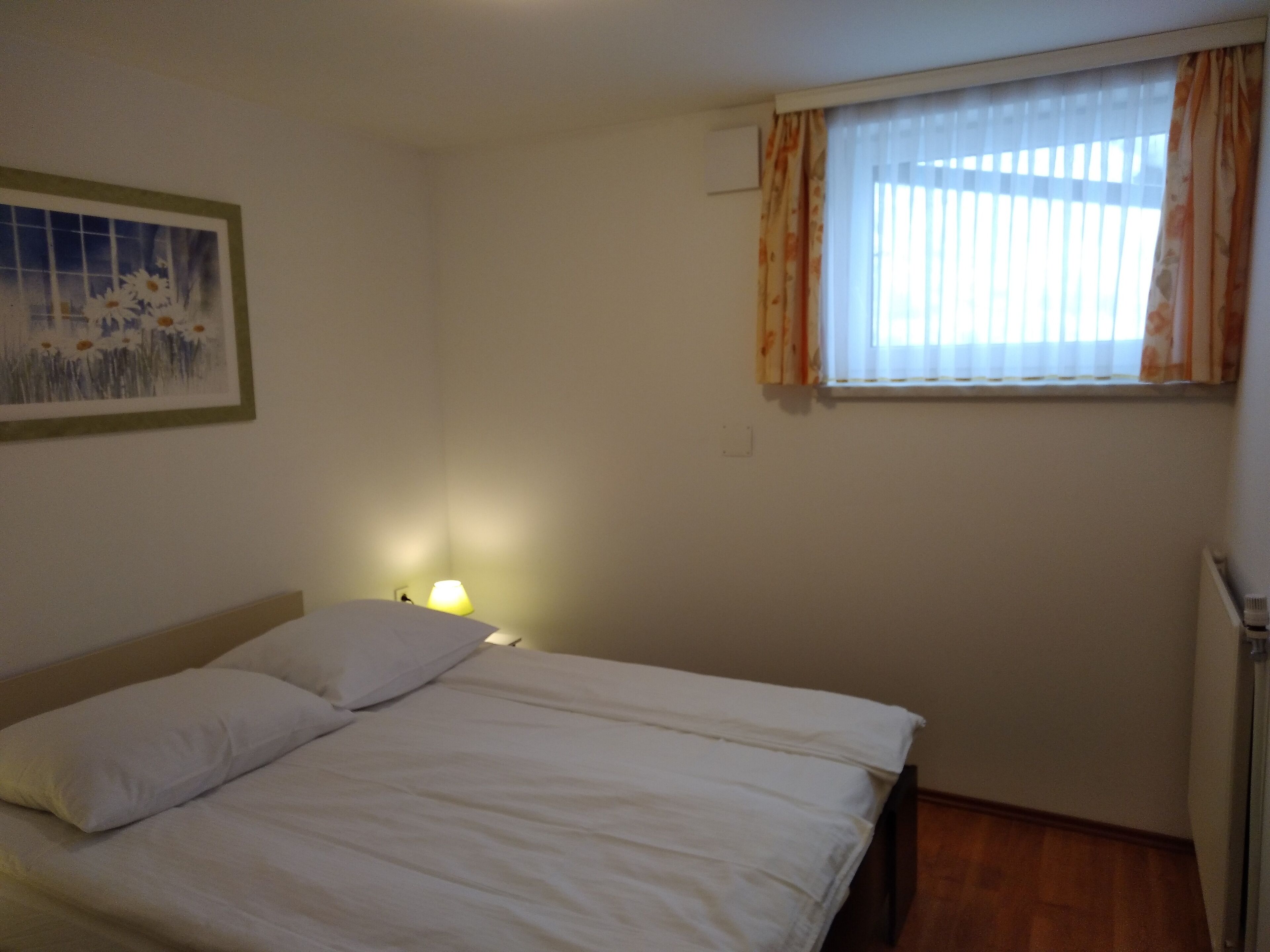 A bedroom with a bed, a nightstand, a lamp, a window with curtains, and a painting on the wall.