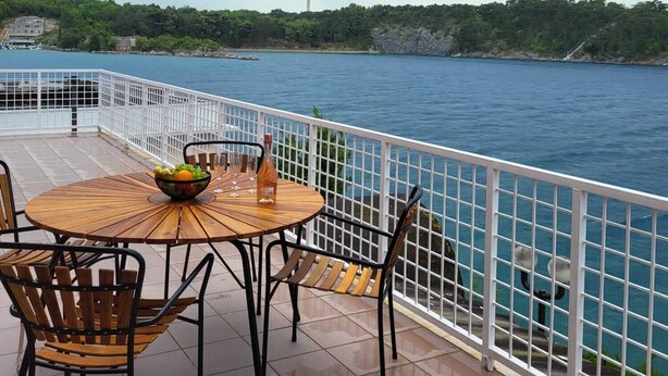 A round wooden table with chairs and a bowl of fruit, overlooking a body of water with a white railing. - Villa Riva (Omisalj)