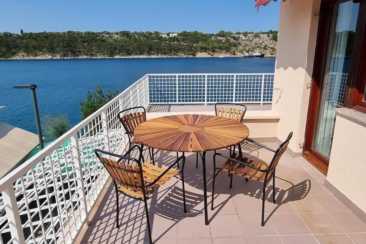 A balcony with a round wooden table and four chairs, overlooking a body of water and a hilly landscape.