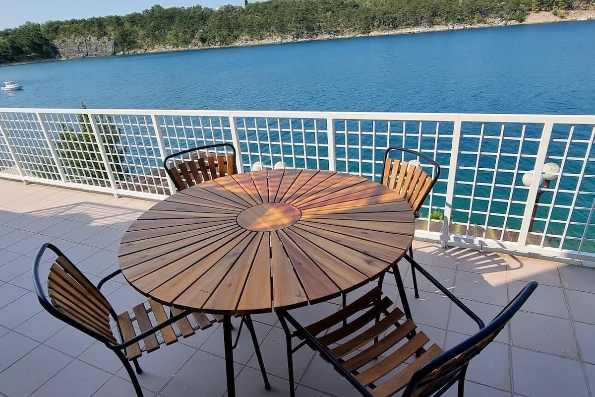 A round wooden table with metal chairs on a tiled patio overlooking a body of water.