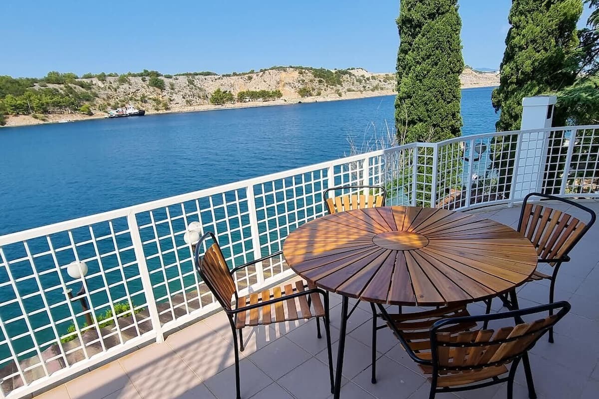 A round wooden table with chairs on a balcony overlooking a body of water.