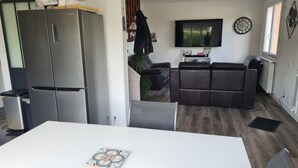 A modern kitchen with a stainless steel refrigerator, a dining table with a decorative plate, a black leather sofa, and a flat-screen TV mounted on the wall. - Maison Dans Lotissement Très Calme 300 Mètres du Centre Ville .10 km de la mer (La Haye)